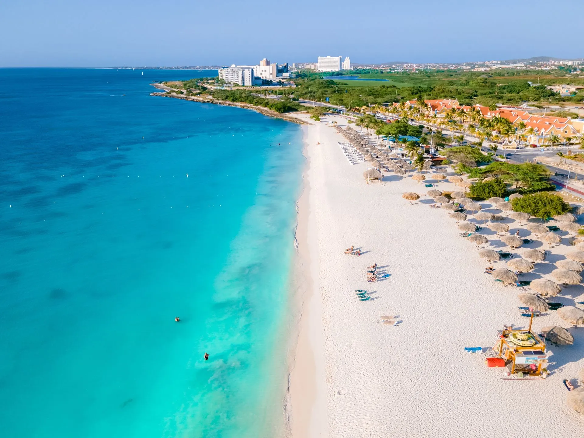 Best Beaches in Aruba – White Sands & Turquoise Seas Wide aerial view of a pristine white-sand beach in Aruba with calm turquoise Caribbean waters and palapas along the shore