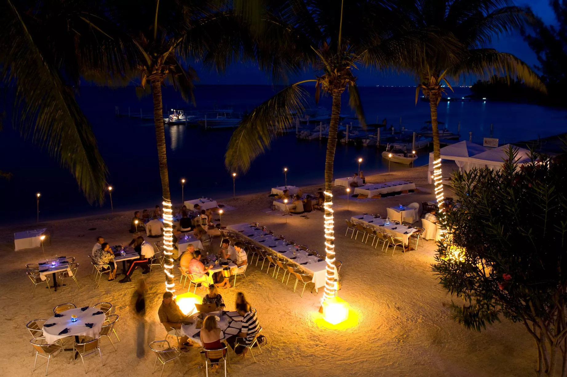 Cayman Islands Nightlife – Beach Bars, Lounges and Evening Experiences Beachfront bar and outdoor dinner setting in Grand Cayman at night with palm trees wrapped in lights and a calm marina in the background