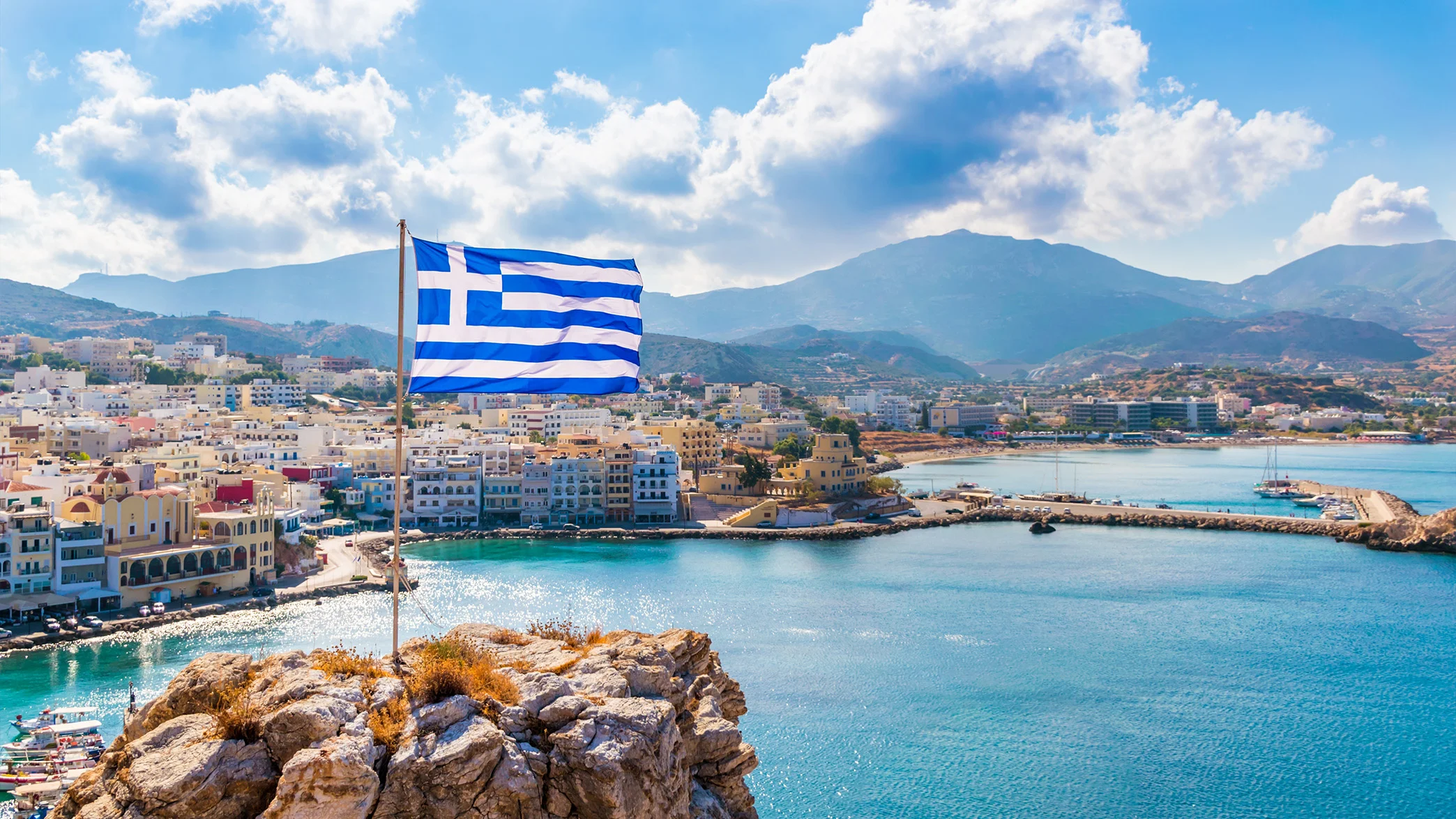 Greece Fast Facts – Greek Flag and Coastal Town Greek flag overlooking a coastal town and harbor in Greece